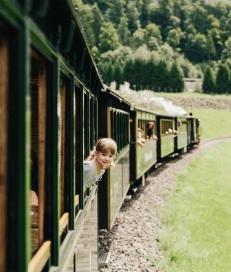 Mädchen schaut lächelnd aus dem Fenster des Wälderbähnle, der historischen Museumsbahn im Bregenzerwald, vorne zieht die Dampflok durch grüne Wiesen.