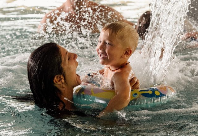 Mutter hält ihr lachendes Kleinkind mit Schwimmreifen unter dem Wasserfall im Hallenbad, dahinter schwimmt der Vater.